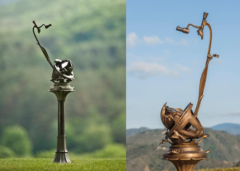 A bronze art sculpture with an abstract, twisting design stands on a tall pedestal. One side features a Porter Garden Telescope against a blue sky backdrop, while the other side displays it on a grassy landscape. The sculpture intricately weaves spherical elements into its form.