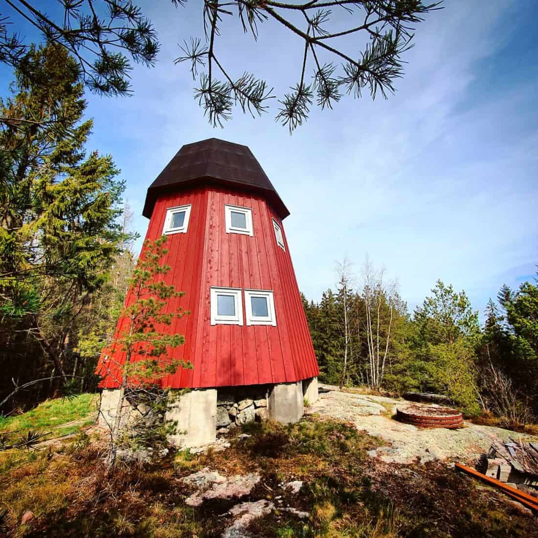 A small, red, tower-like cabin reminiscent of a Vann Hotel in Sweden stands on a rocky clearing surrounded by trees under a clear sky. The structure boasts a dark roof and several square windows. Branches naturally frame the scene.