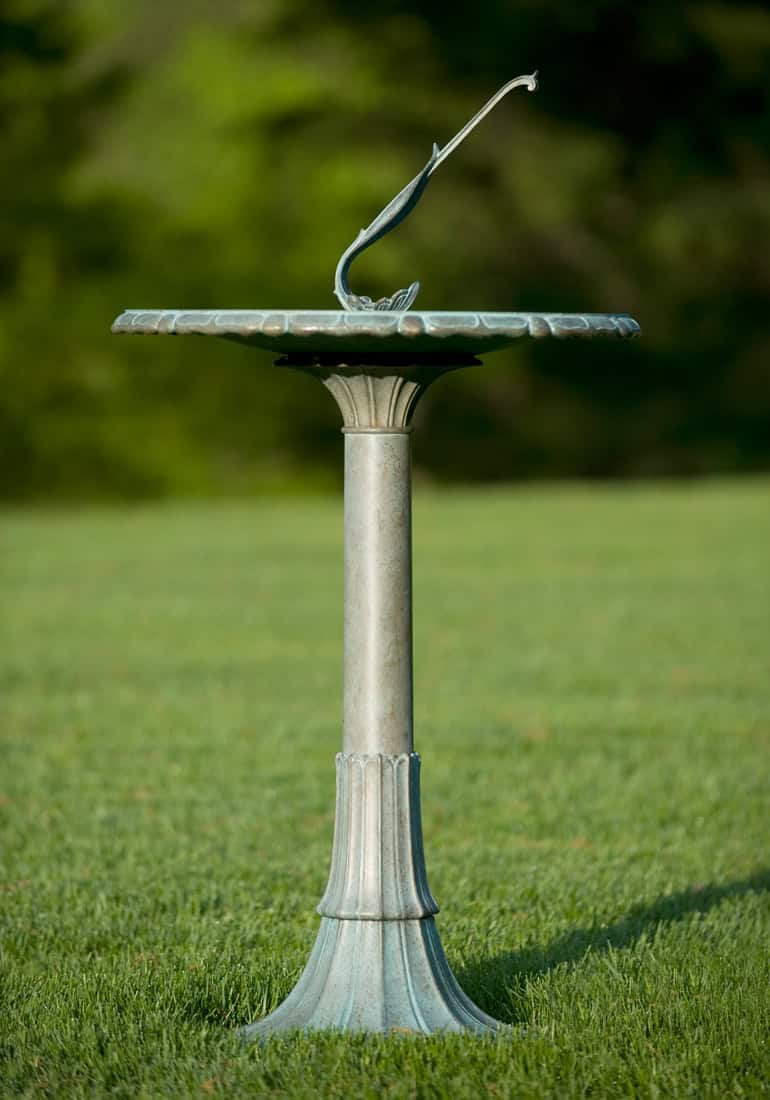 A metal sundial stands on a grassy garden lawn, with a vertical gnomon casting a shadow on the circular dial. The blurred greenery in the background highlights the sundial and its intricate design, much like an intricately crafted telescope highlighting celestial wonders.