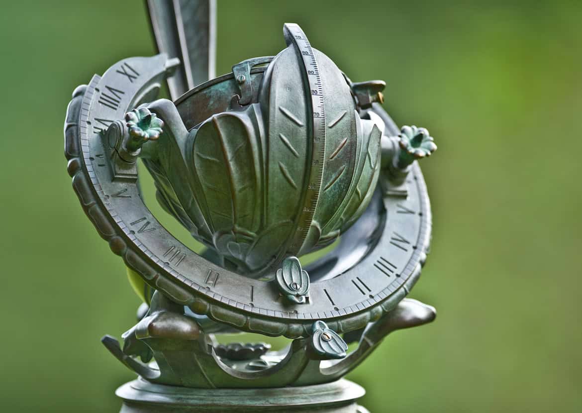 A metallic armillary sundial with Roman numerals, featuring an intricate design of rings and arrows, stands elegantly in the garden on a blurred green background.