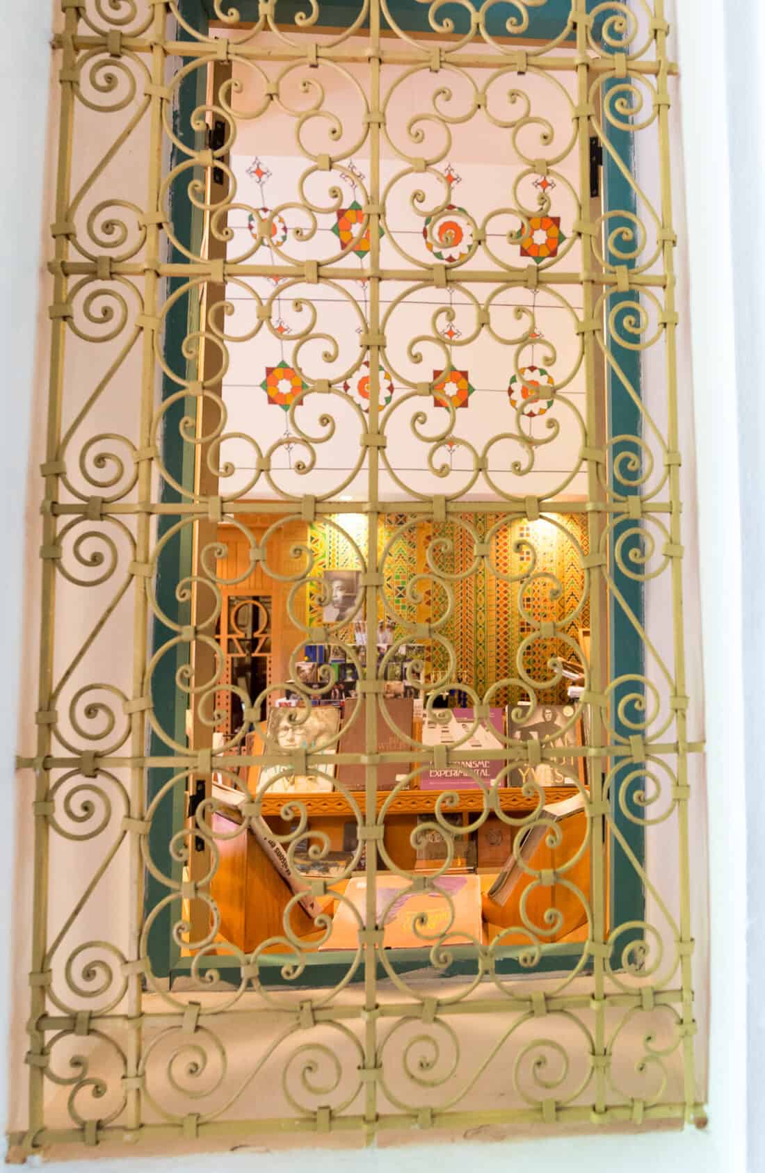 An ornate metal grille with floral patterns, reminiscent of the Majorelle Garden, in front of a window, behind which a person is partially visible.