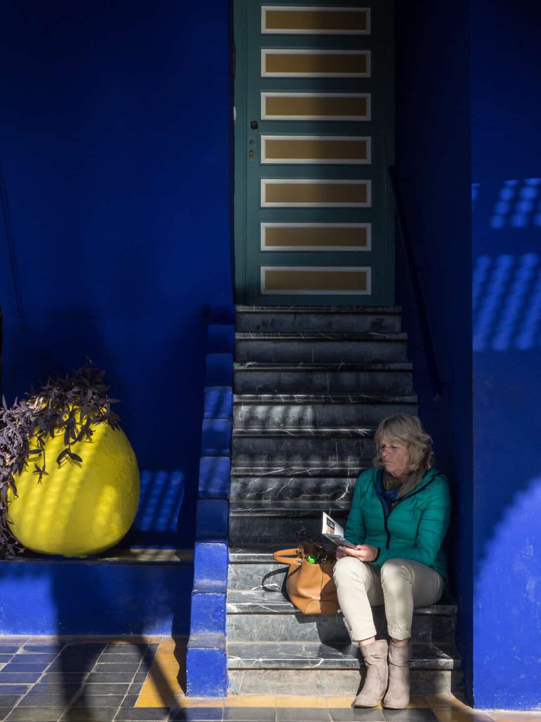 A woman sitting on steps reading a brochure beside a bright yellow pot with plants in Majorelle Garden.