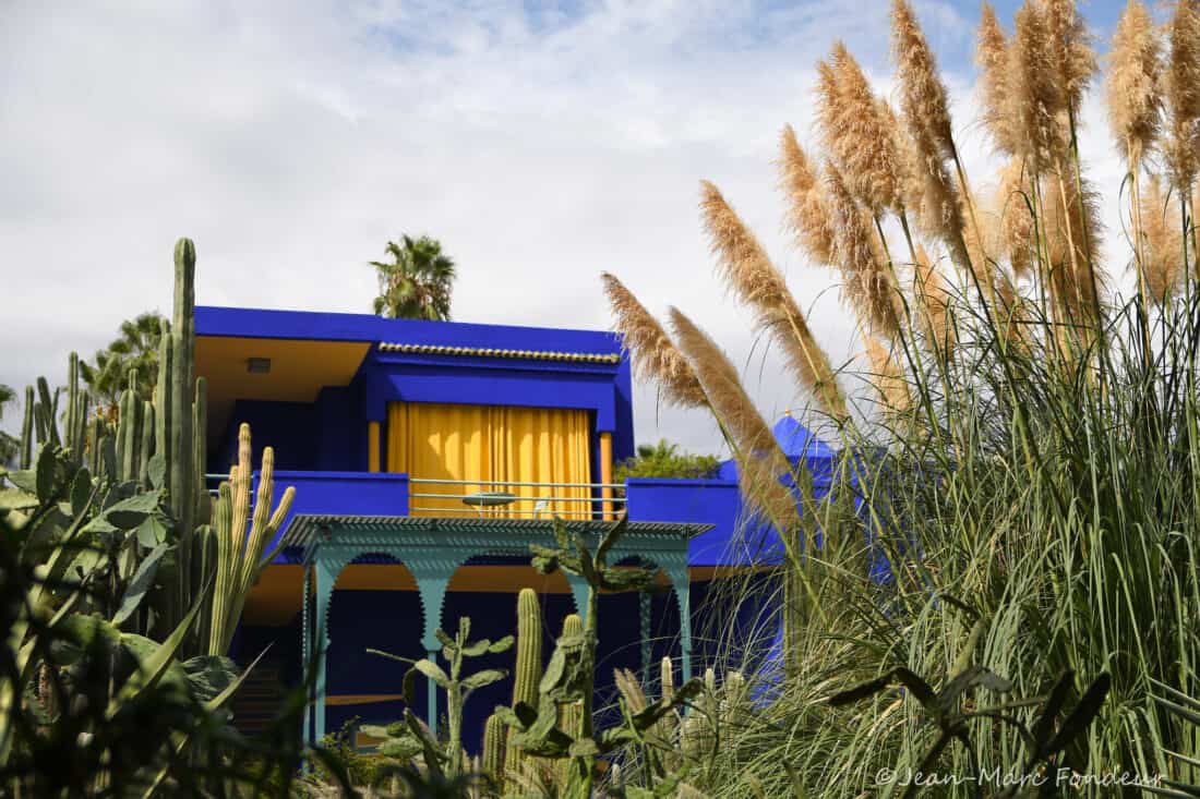 A vibrant blue building with yellow accents in Majorelle Garden, partially obscured by lush greenery under a cloudy sky.