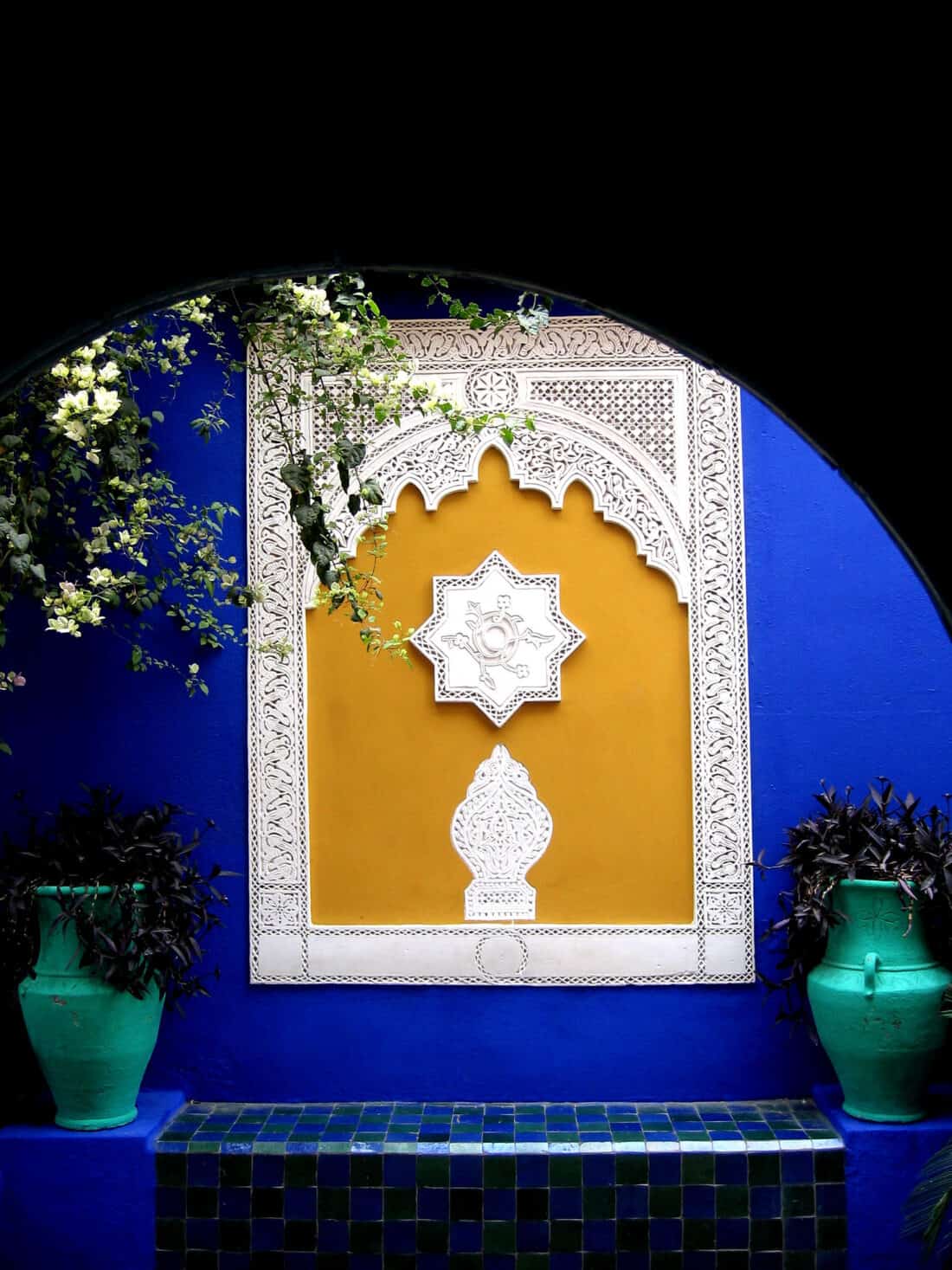 An ornate window with traditional Moroccan design, framed by vibrant blue walls of Majorelle Garden and flanked by potted plants.