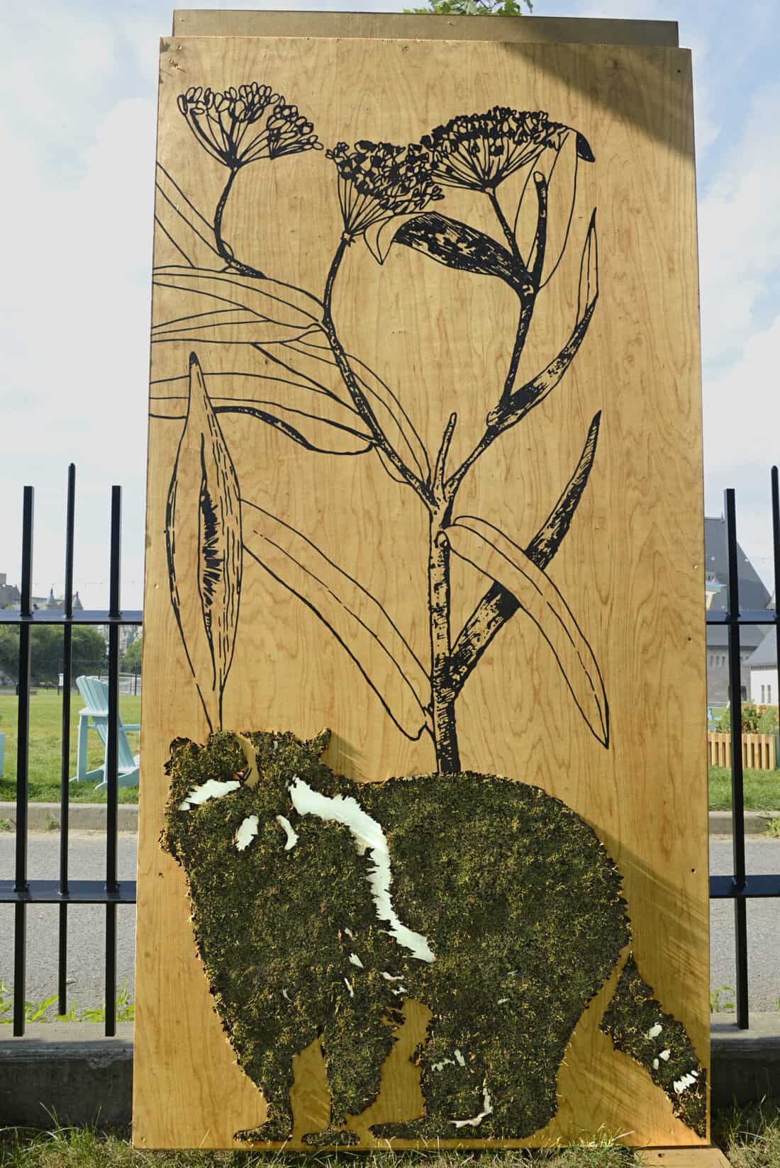 A wooden panel art piece by Edina Todoki showcases a silhouette of a red panda crafted with moss. Above it, delicate black line drawings of tall flowers and leaves add elegance. Displayed outdoors, this exquisite vertical garden art complements the fence and grassy area in the background.