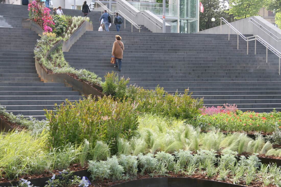 A person walks up wide, gray stone steps with lush green and flowering plants lining the path, resembling a flowing garden. The scene is set in an urban area of Bilbao, Spain, with modern architecture and people in the distance.