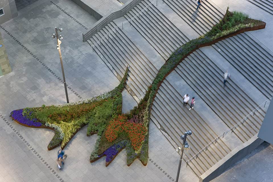 Aerial view of a large, abstract plant sculpture with vibrant flowers and greenery set between stairways in Bilbao. This flowing garden attracts visitors moving around the sculpture on a paved plaza, providing a sense of scale and perspective.