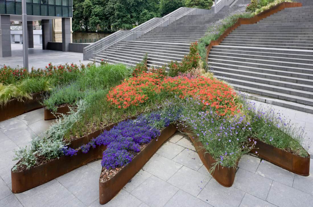 The urban landscape of Bilbao features star-shaped metal planters filled with vibrant red, purple, and green plants. These flowing garden installations rest on a paved surface beside wide concrete stairs, framed by trees and the sleek architecture of modern buildings in the background.