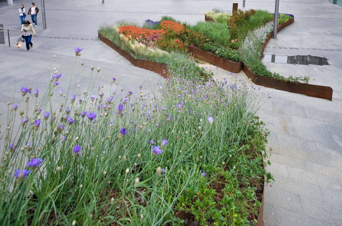 A paved urban area in Bilbao showcases vibrant flower beds, with purple and red blossoms bordered by rust-colored metal edges. Three people walk along the wide path of this flowing garden, adding a sense of scale to the spacious setting.