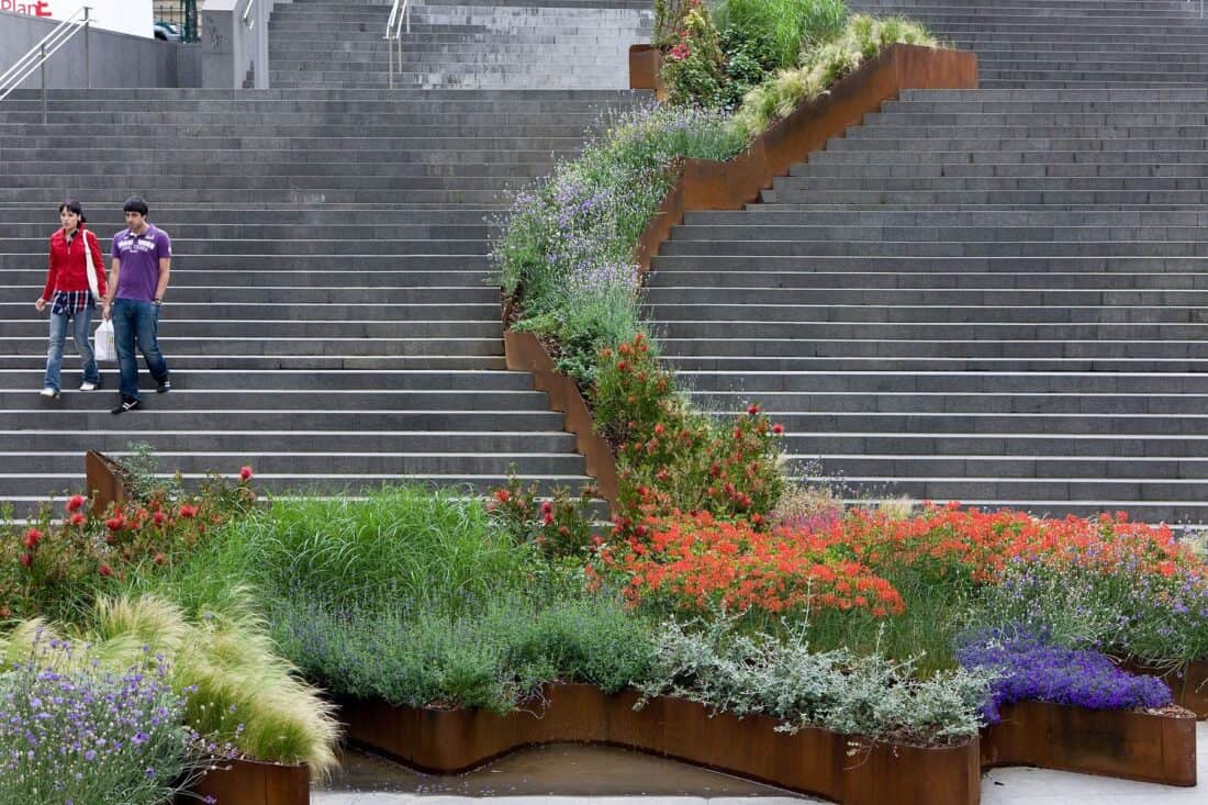 In Bilbao, two people walk down a large stone staircase that features a flowing garden with lush greenery and vibrant flowers. The stairs create a striking contrast with the colorful plants, adding to Spain's enchanting charm.