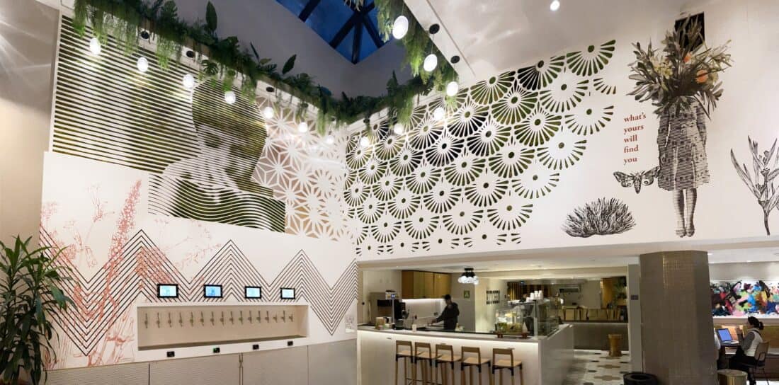 A modern café interior with artistic wall designs, including abstract patterns and Vertical Garden Art featuring a woman holding flowers above her head. There are hanging plants, taps with electronic screens, bar stools, and a counter with staff.