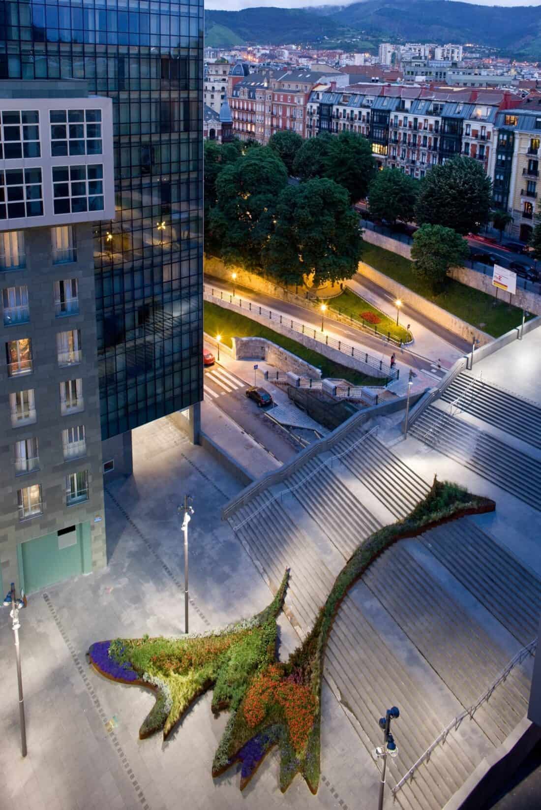 Aerial view of Bilbao's modern urban landscape with a large, flowing garden shaped like an abstract flower next to a staircase. Surrounding buildings and streetlights are visible, with a backdrop of trees and distant hills under the twilight sky in Spain.