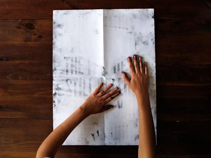 A person with dark-painted nails gets their hands dirty, spreading them on a large, white, slightly dirty sheet of paper with faint, unreadable text, set on a dark wooden surface.