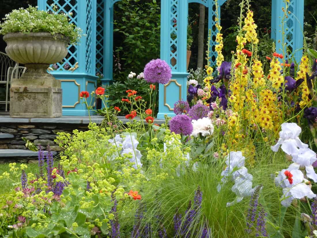 A vibrant garden scene reminiscent of the 2010 Chelsea Flower Show features various colorful flowers, including purple alliums, yellow foxgloves, and red poppies, in front of a blue gazebo. A stone planter with lush greenery enhances the stunning plant combos on display.