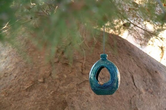 A teal ceramic birdhouse with an oval-shaped design hangs gracefully from a tree branch in the garden. In the background, a large rock is framed by blurred greenery, reminiscent of the serene landscapes near Joshua Tree.