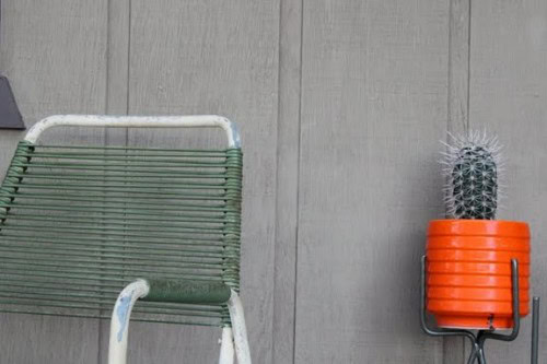 A green, vintage metal chair is placed against a plain wooden wall, evoking the rustic charm of a Joshua Tree garden. Beside it, a small cactus sits in a bright orange planter, adding to the simplicity and serenity of this outdoor setting.