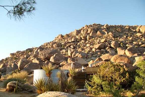 A desert landscape with a rocky hill under a clear blue sky. In the foreground, a Stock Tank Pool and a wooden structure surrounded by sparse vegetation, including bushes and a few small trees reminiscent of those in Joshua Tree, are visible.