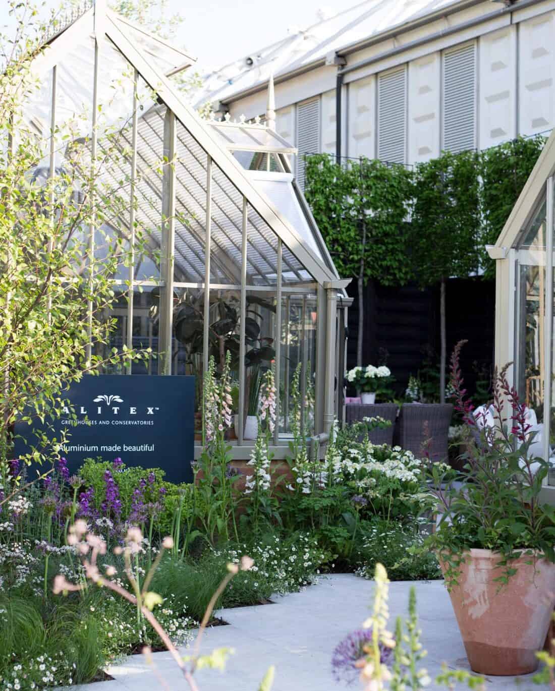 A charming garden scene featuring a glass greenhouse with a sign reading "Alitex: Aluminium made beautiful." The pathway, reminiscent of Marston & Langinger's designs, is bordered by lush greenery, flowers, and potted plants. A seating area with a table and chairs is visible in the background.