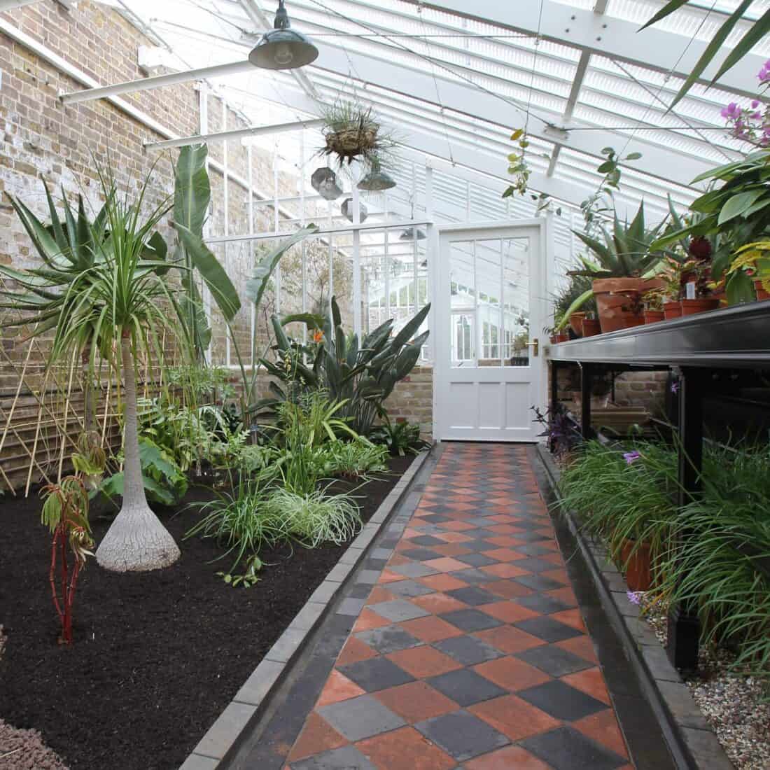 A bright greenhouse by Marston & Langinger features a tiled walkway and a variety of tropical plants. Plants are in the ground and in pots placed on metal shelves. A white door is at the end of the greenhouse, with large windows and a brick wall on one side. Hanging plants adorn the ceiling.