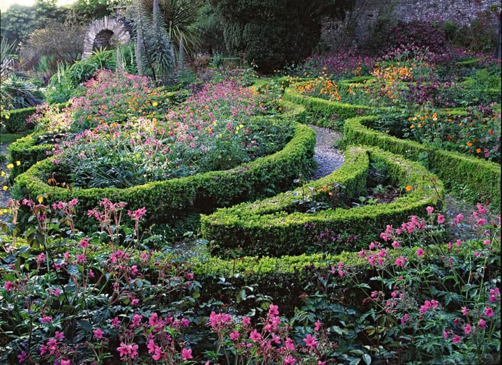 A vibrant garden with winding paths lined by neatly trimmed hedges draws you close. The garden, reminiscent of an Allan Pollok-Morris photograph, bursts with colorful flowers in pinks, oranges, and purples. In the background, a stone archway and lush greenery are visible, inviting further exploration.