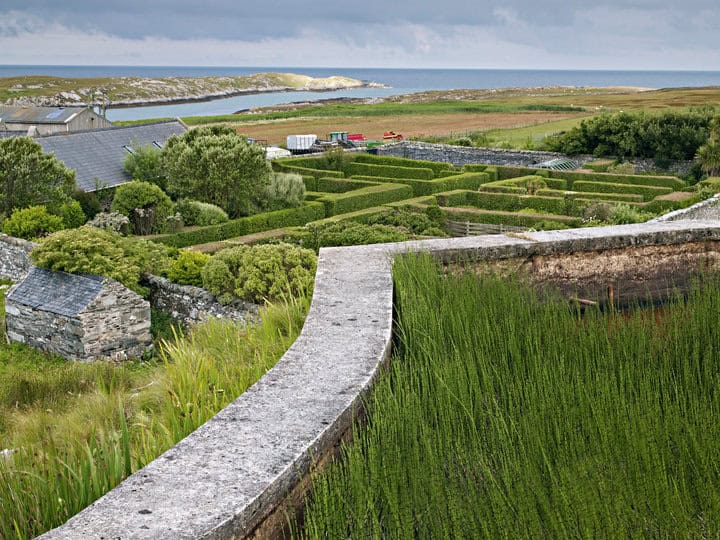 A lush garden with neatly trimmed hedges and a stone wall overlooks a scenic coastline, reminiscent of Allan Pollok-Morris's vivid imagery. The overcast sky casts a gentle mood as the landscape stretches to a distant view of the sea and rocky headlands, inviting contemplation akin to an evocative book review.