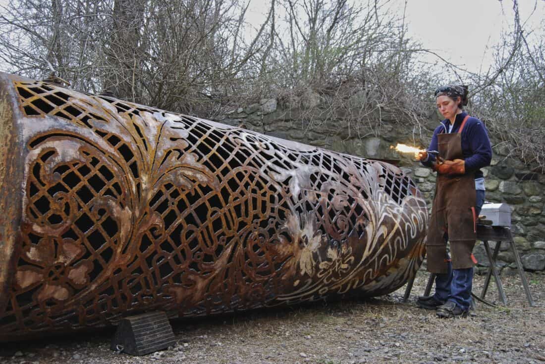 A person in work attire uses a torch on a large, intricately designed metal cylinder, reminiscent of Cal Lane's signature style. The cylinder features ornate floral patterns. Trees and a stone wall are in the background, contrasting with the grass burning gently nearby.