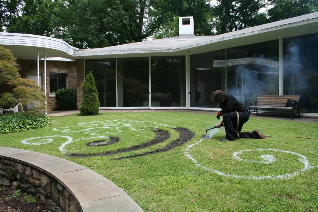 A person is creating swirling designs on a grassy lawn using white and dark materials, reminiscent of Cal Lane's artistic style. In front of a modern house with large windows and a chimney, the scene is alive with smoke rising from grass burning where the intricate patterns take shape.