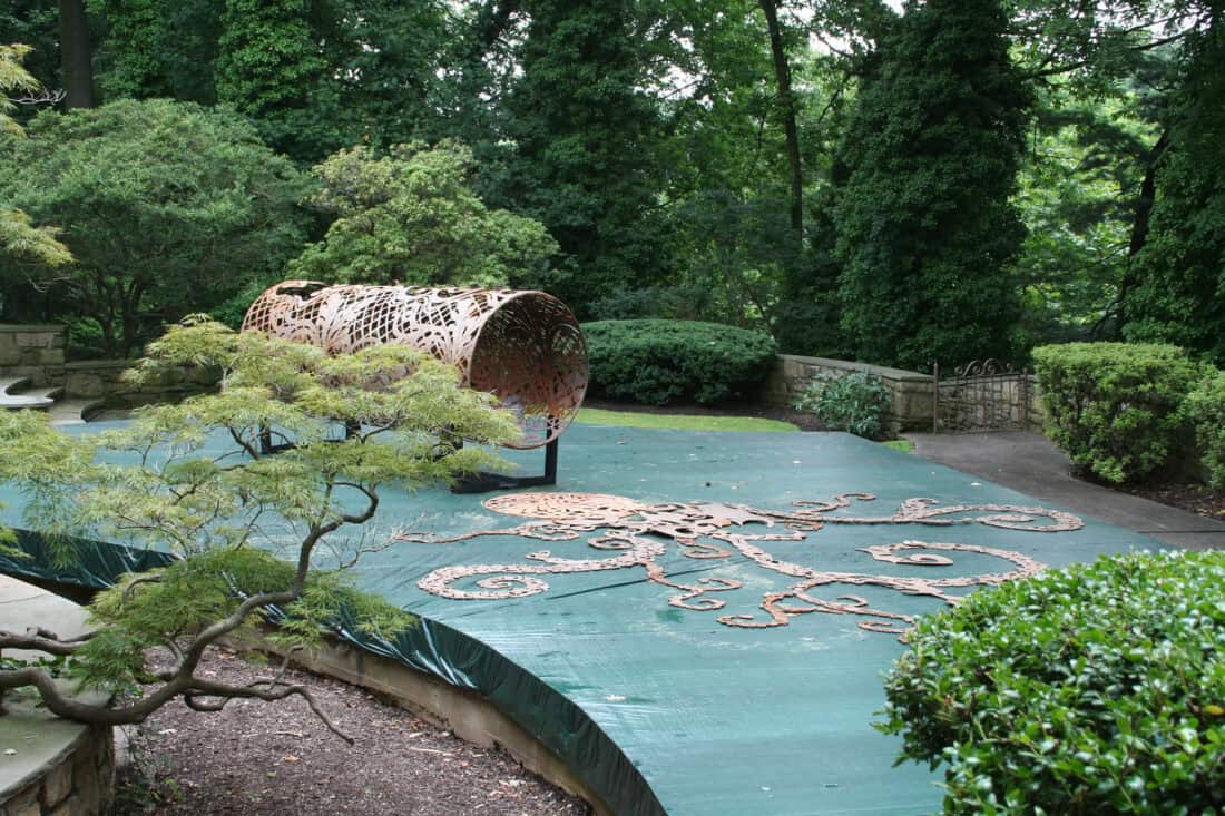 Inspired by Cal Lane, this sculpture art installation transforms a lush garden setting. A cylindrical, intricately patterned metal structure stands on a large, flat, green platform surrounded by trees and bushes, with an abstract design reminiscent of grass burning etched into the platform surface.