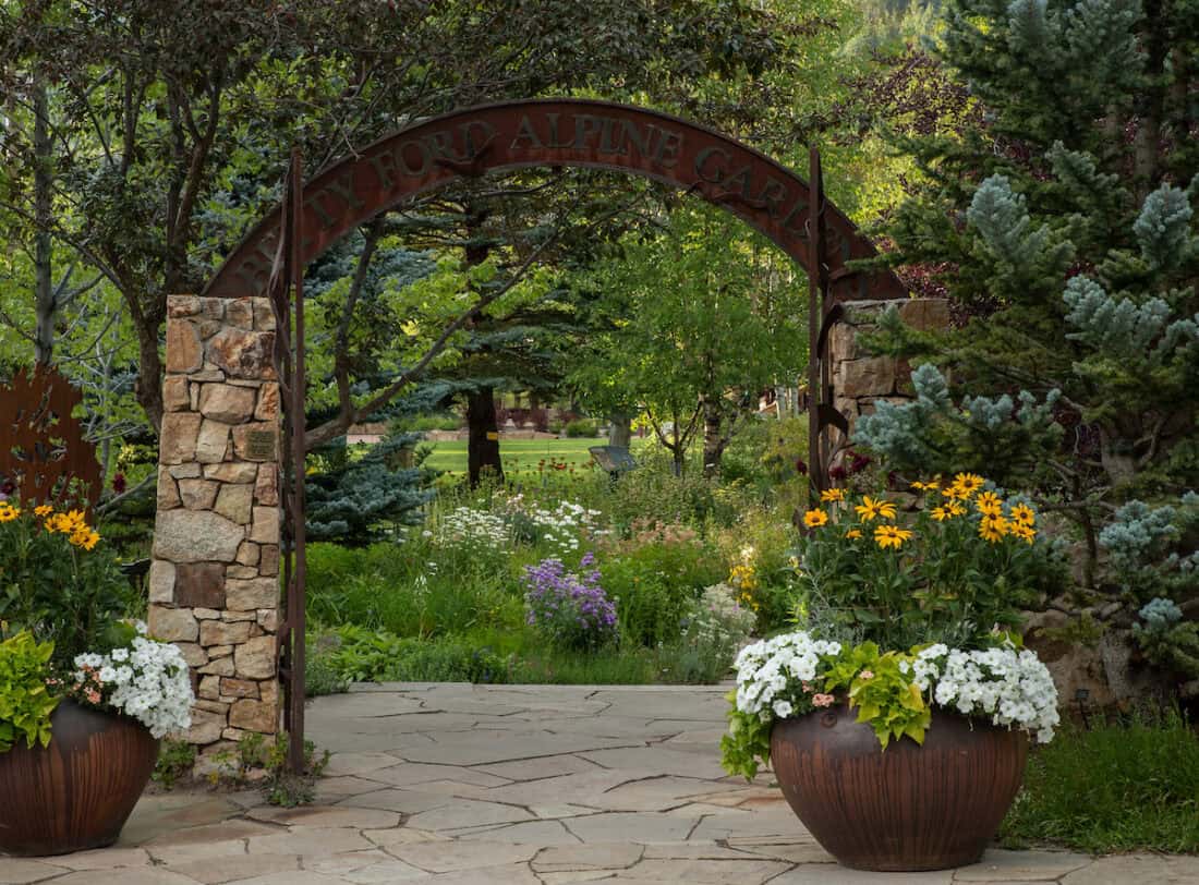 A stone pathway leads through an arched entrance to a lush garden brimming with vibrant flowers and towering trees in Vail, Colorado. The sign reads "Betty Ford Alpine Gardens." Large planters with yellow and white blooms elegantly frame the entrance.