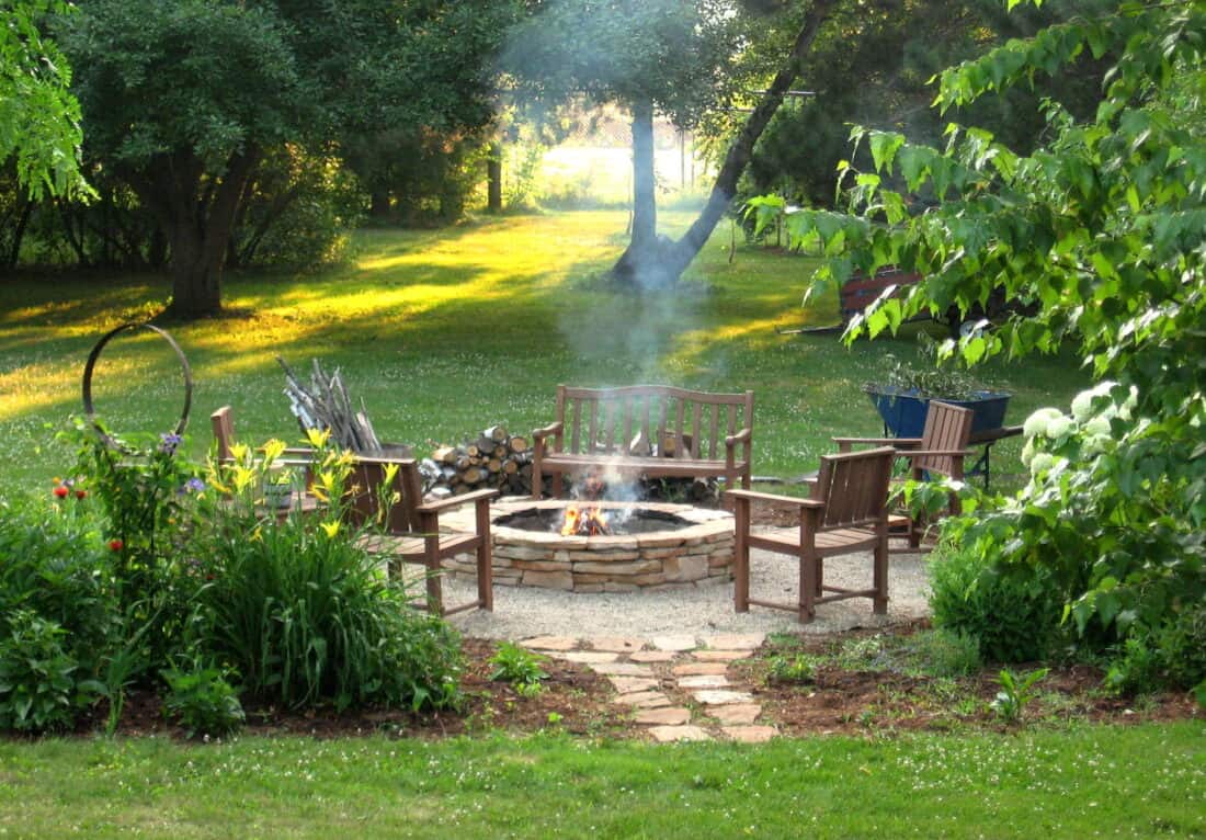 Erin's Fire Pit is the centerpiece of a serene backyard, with four wooden chairs surrounding it on a gravel circle. The area is bordered by greenery, plants, and tall trees. Smoke gently rises from the fire pit as sunlight filters through the trees, creating a peaceful ambiance.