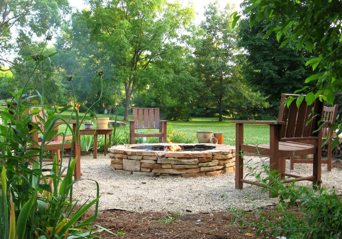 A serene garden setting featuring Erin's Fire Pit, surrounded by wooden chairs and lush greenery. The fire pit rests on a gravel base, with potted plants adding to the natural ambiance. Trees and a grassy area are visible in the background, completing this Before & After transformation.