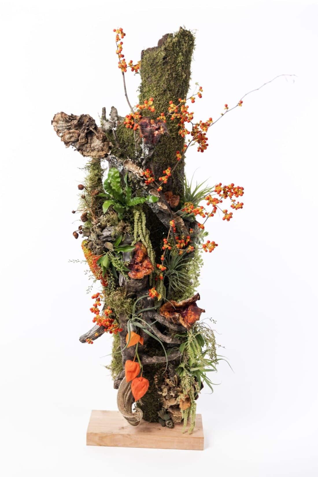 A vertical composition of assorted natural elements from a woodland garden, including moss, wood, and orange berries, arranged on a wooden base to form a decorative piece.