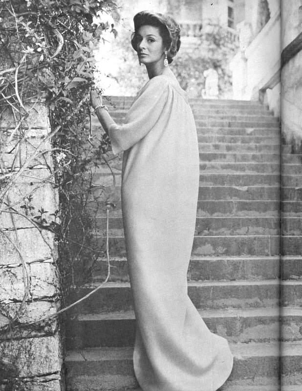 A woman in a long, elegant gown stands on outdoor stone steps, reminiscent of Marella Agnelli's style, looking back towards the camera. The black-and-white scene captures her amidst lush greenery and an old stone structure, evoking the tranquility of a Marrakech Garden.