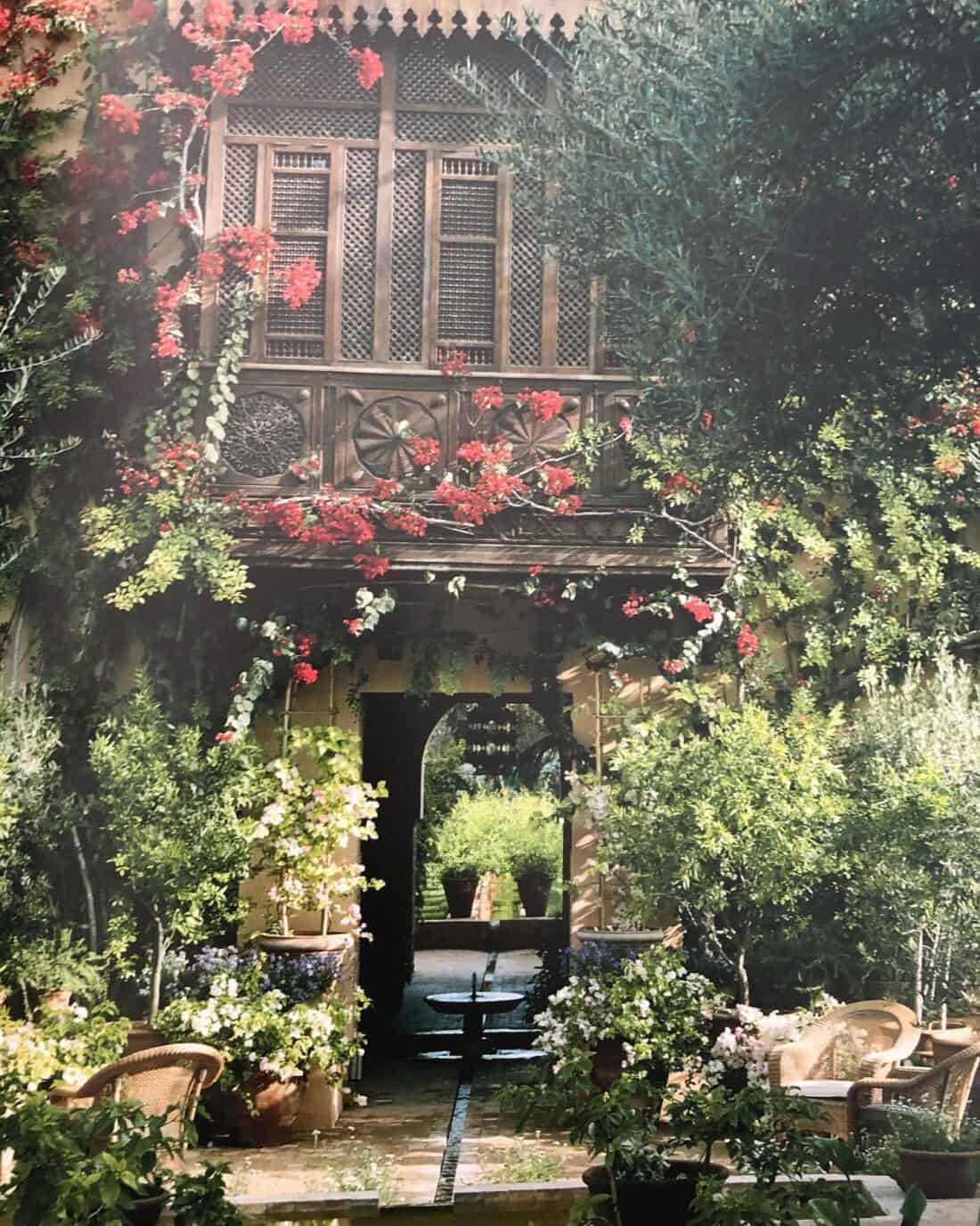 A charming Marrakech Garden features lush greenery and vibrant red flowers climbing a rustic wooden balcony. The scene includes a table and chairs surrounded by potted plants, leading to an arched doorway with more plants visible inside, reminiscent of a serene garden gallery.