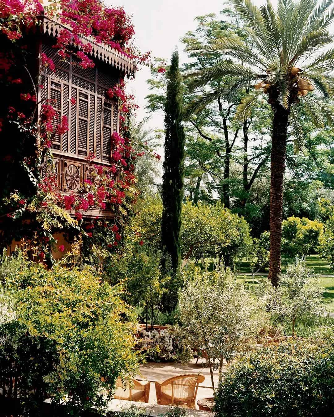 A lush Marrakech Garden features a tall palm tree, various shrubs, and a vine-covered building with intricate wooden architecture reminiscent of Marella Agnelli's style. A round patio table with wicker chairs is set in the foreground under a bright sky.