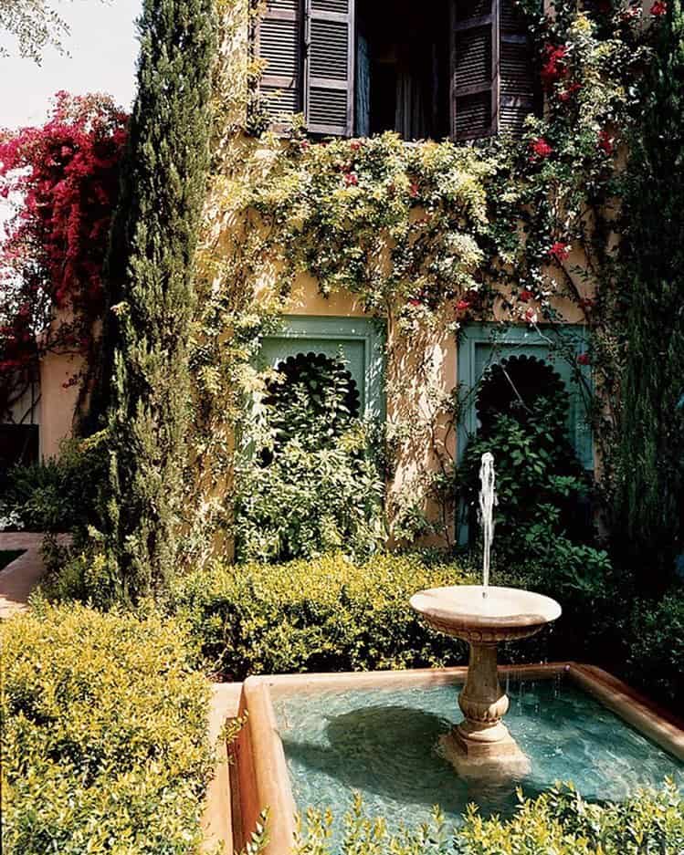 A tranquil Marrakech garden scene with a small fountain in the foreground. The two-story building is draped in green vines and red flowers, reminiscent of a living Garden Gallery. Tall, narrow trees frame the windows with dark wooden shutters, while lush green hedges complete the serene tableau.