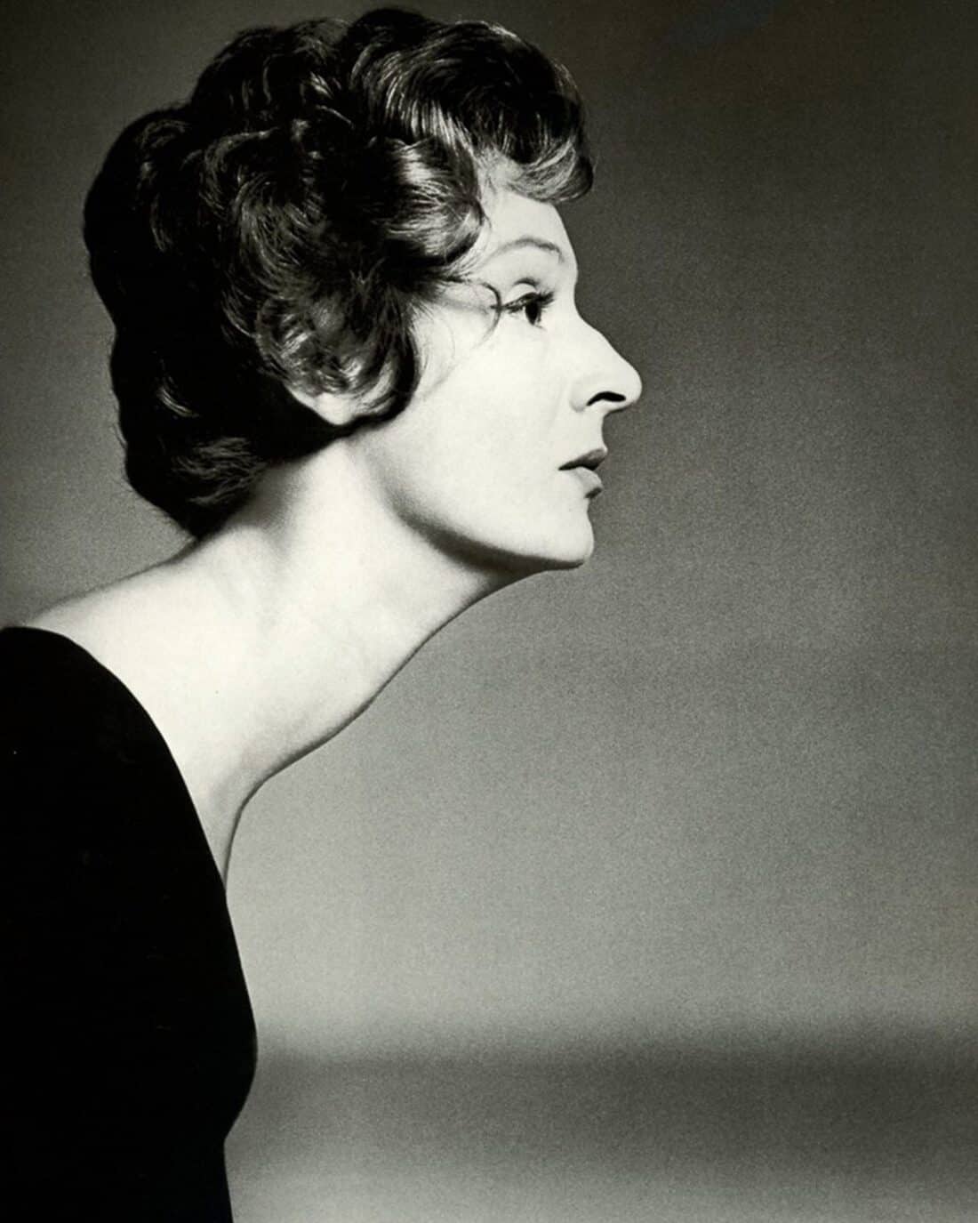 A black-and-white portrait reminiscent of Marella Agnelli, capturing a person in side profile with a short, curly hairstyle. They wear a dark top against a gradient background from light to dark gray, akin to the shifting hues of a Marrakech garden. The lighting enhances their facial features elegantly.