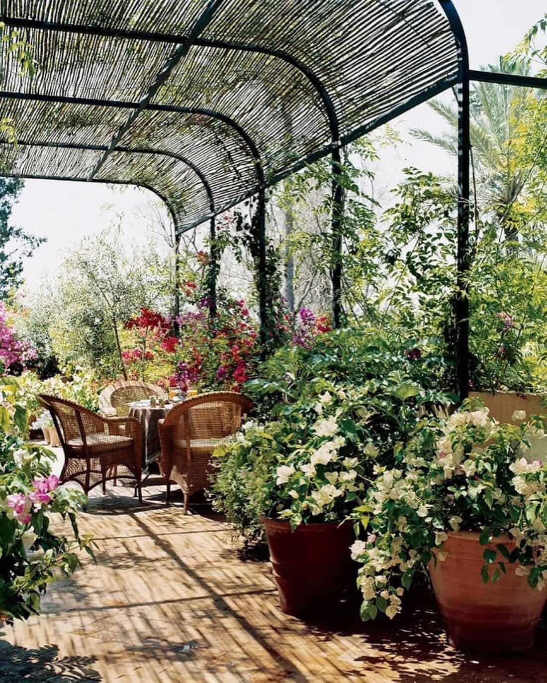 A sunlit garden patio features wicker chairs around a small table, surrounded by lush potted plants with vibrant flowers reminiscent of a Marrakech Garden. A bamboo pergola overhead provides shade, creating a peaceful and inviting atmosphere, like strolling through a serene garden gallery.