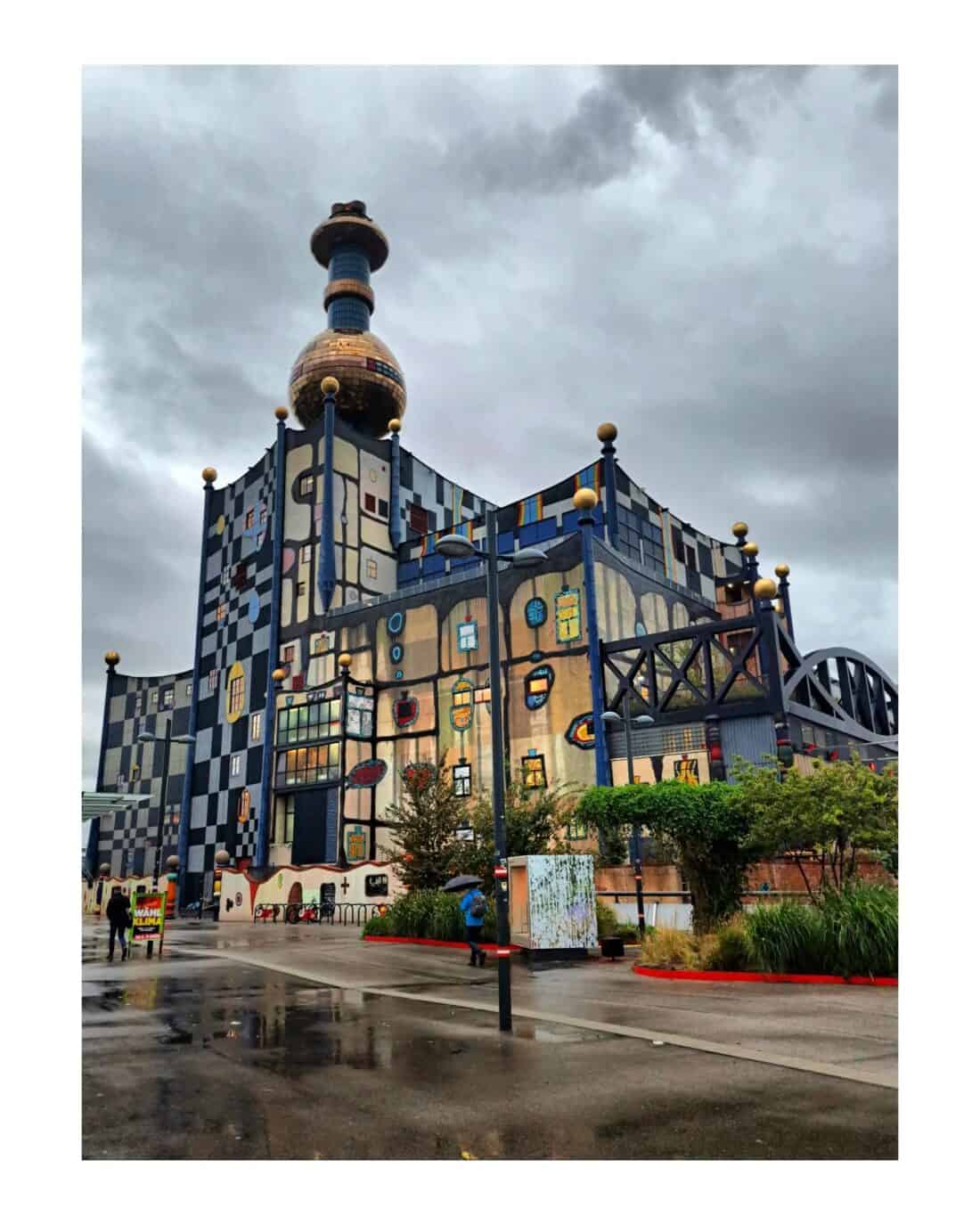 The building, reminiscent of Friedensreich Hundertwasser's work, showcases colorful, irregular patterns and a unique golden dome against a cloudy sky. The facade features curved lines and small, round windows. Greenery and wet pavement in the foreground suggest recent rain.