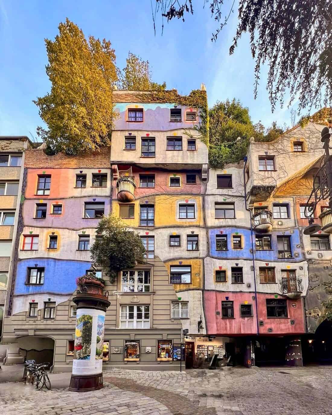 A colorful, whimsical multi-story building with uneven windows and unique balconies, reminiscent of Friedensreich Hundertwasser's style, is surrounded by trees. The structure boasts a patchwork of pastel colors with a kiosk in front, all set against a clear blue sky.