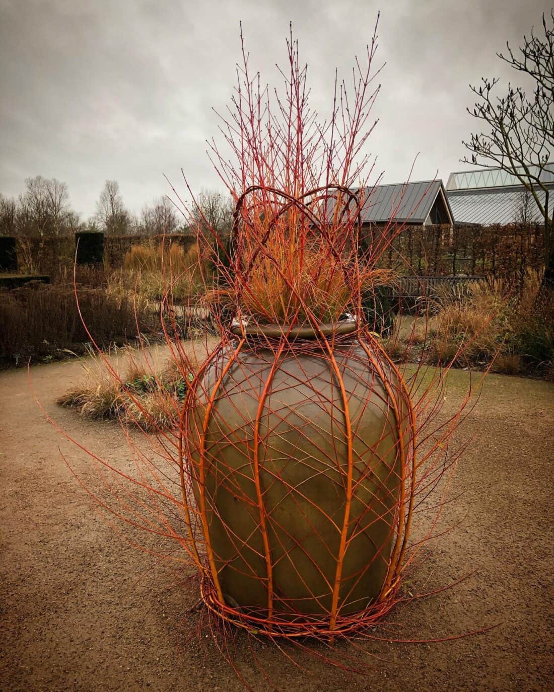 A large, earthy-colored ceramic pot is adorned with vibrant red and orange branches that resemble dancing flames, set on a garden pathway. In the background, trees and shrubs surround two wooden shed-like structures under an overcast sky.