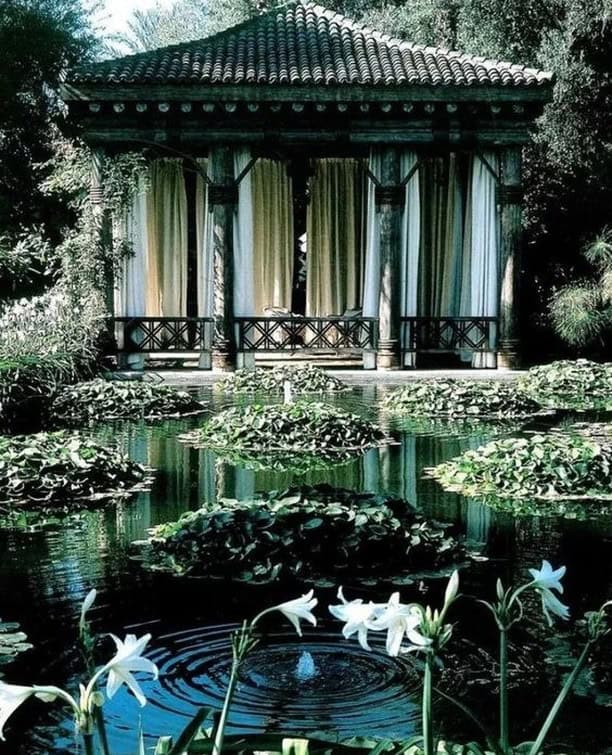 A serene pond with white lilies and lush green leaves sits in front of a pavilion adorned with curtains, reminiscent of a Marrakech Garden. Surrounded by trees and foliage, the water reflects the greenery, creating a tranquil, natural scene that could grace any Garden Gallery.