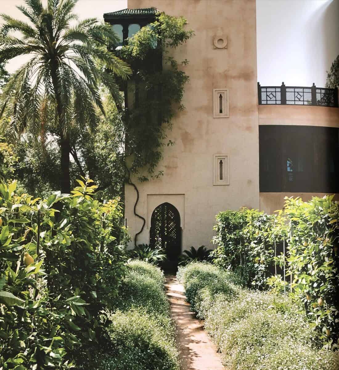 A lush garden pathway leads to a tall, beige building with arched windows and a tower, reminiscent of Marrakech Garden, surrounded by green foliage and palm trees under clear skies.