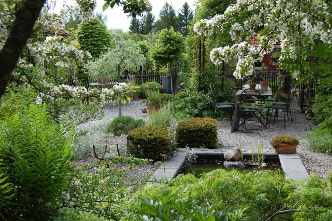 A lush garden gallery features blooming trees and various green plants surrounding a small pond with rocks. In the background, a gravel path leads to Mariana's seating area with a table and chairs, framed by dense foliage and trees.