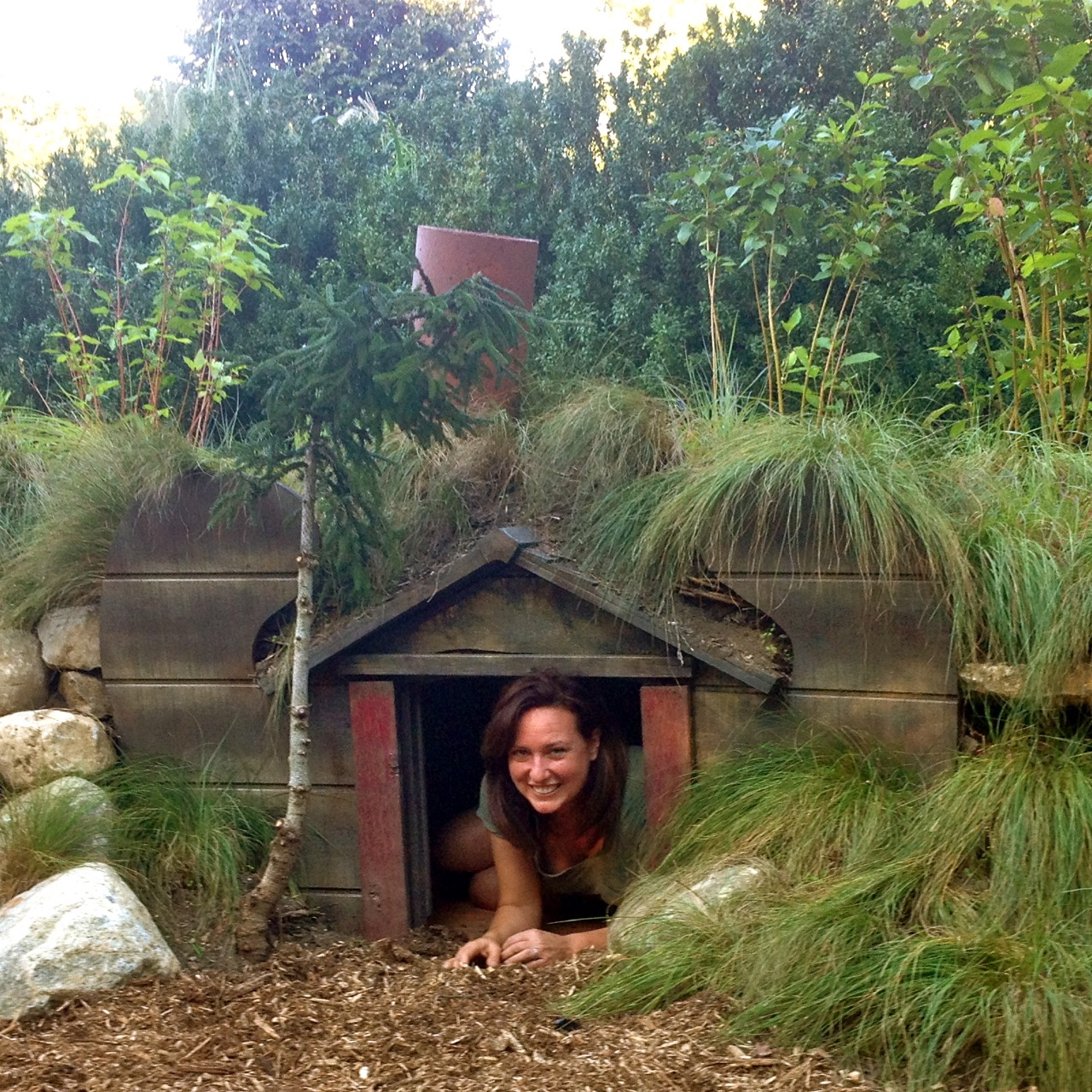 A woman with brown hair is smiling and crawling out of a small, rustic wooden structure that resembles a hobbit house, set into a grassy hillside. Surrounded by the lush greenery of a hobbit garden, she appears to be enjoying the playful, fairy-tale setting.