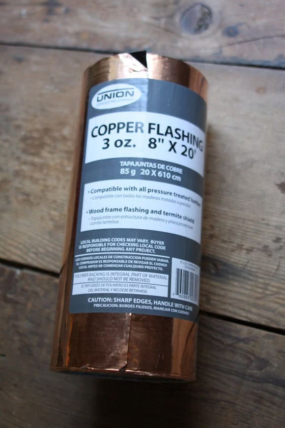 A roll of Union Copper Flashing with dimensions 3 ounces, 8 inches by 20 feet is displayed standing upright on a wooden surface. The label includes compatibility information, warnings, and usage instructions in both English and Spanish, detailing its use to control slugs effectively.