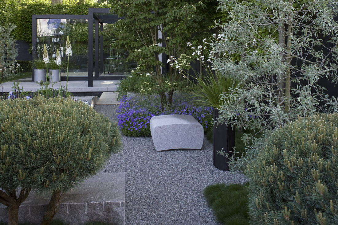 A modern garden, inspired by Ulf Nordfjell, features a gravel path with large square stones surrounded by lush green plants and purple flowers. A stone bench nestles amidst the greenery, while a sleek glass structure stands gracefully in the background.