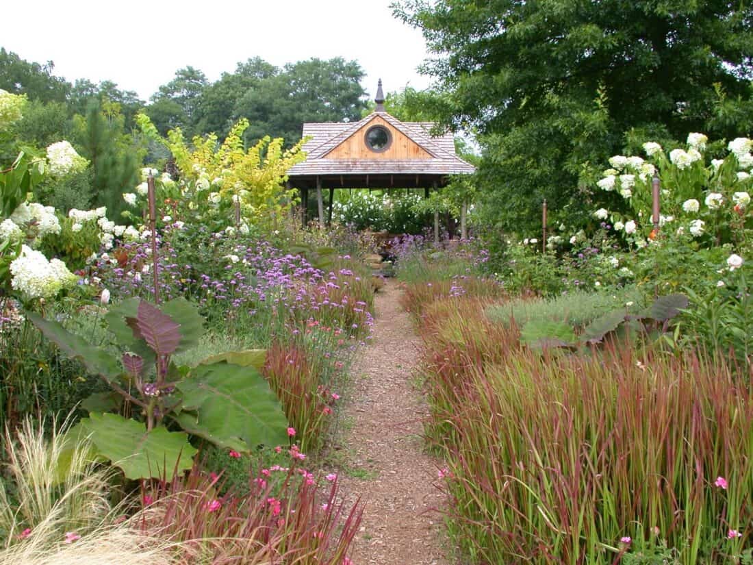 A garden path leads to a wooden gazebo surrounded by lush greenery and colorful flowers, including purple, pink, and white blossoms—like a nursery of nature's love. Tall grasses and large green leaves line the path under a cloudy sky.