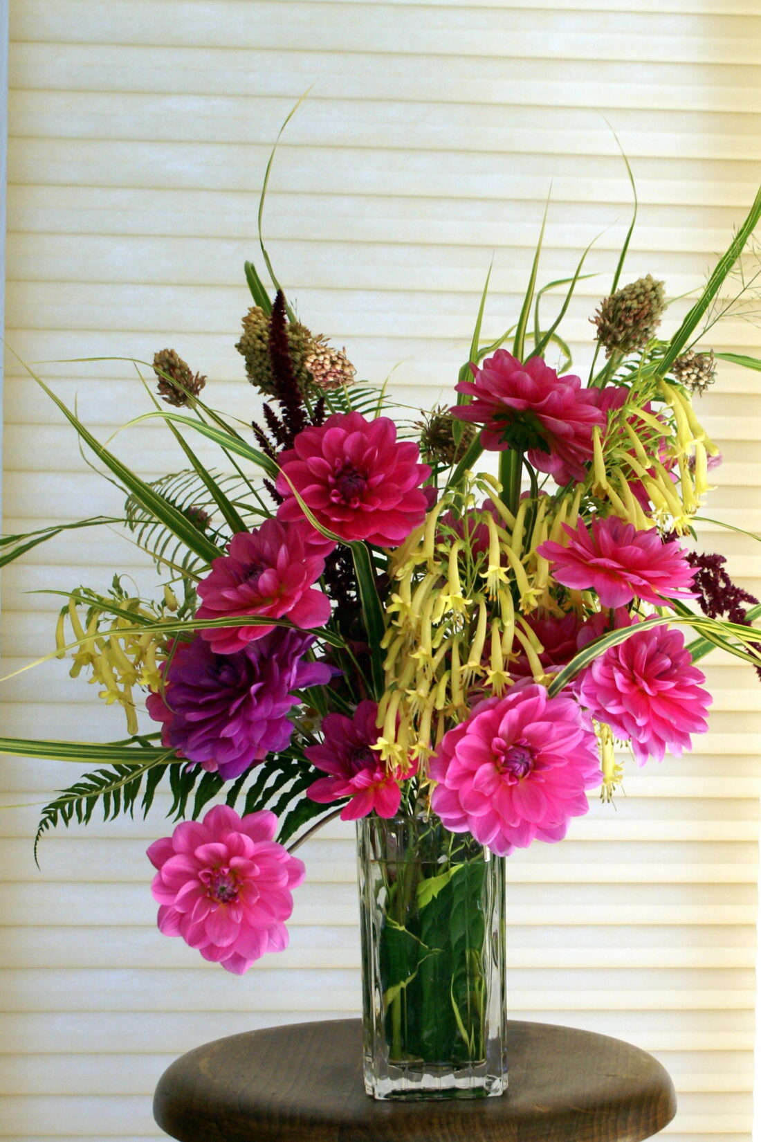 A vibrant floral arrangement in a clear rectangular vase, featuring pink and purple flowers with long green leaves and yellow accents, including Cape Fuchsia blooms, sits atop a round wooden table against a softly textured light background.