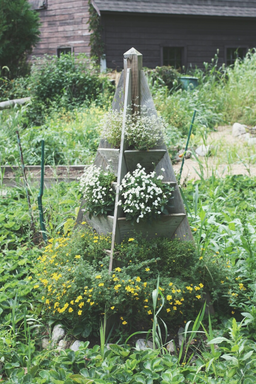 A wooden tiered strawberry tower planter in a pyramid shape stands in a lush garden. The planter has different tiers filled with blooming white and yellow flowers, with greenery all around. An old wooden building can be seen in the background.